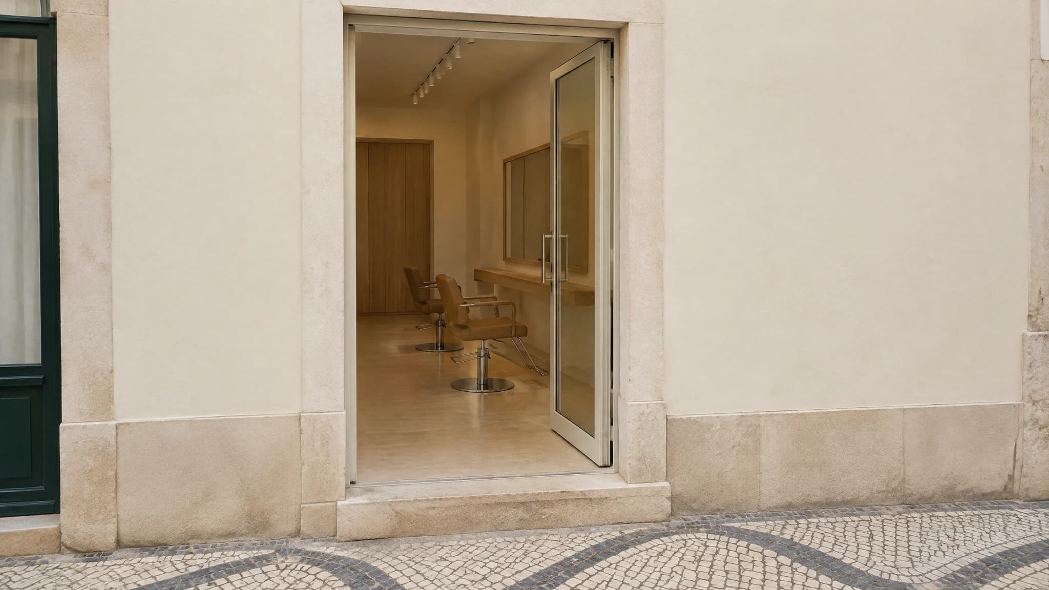 Porta de vidro aberta numa fachada creme, com cadeiras lá dentro e calçada em mosaico