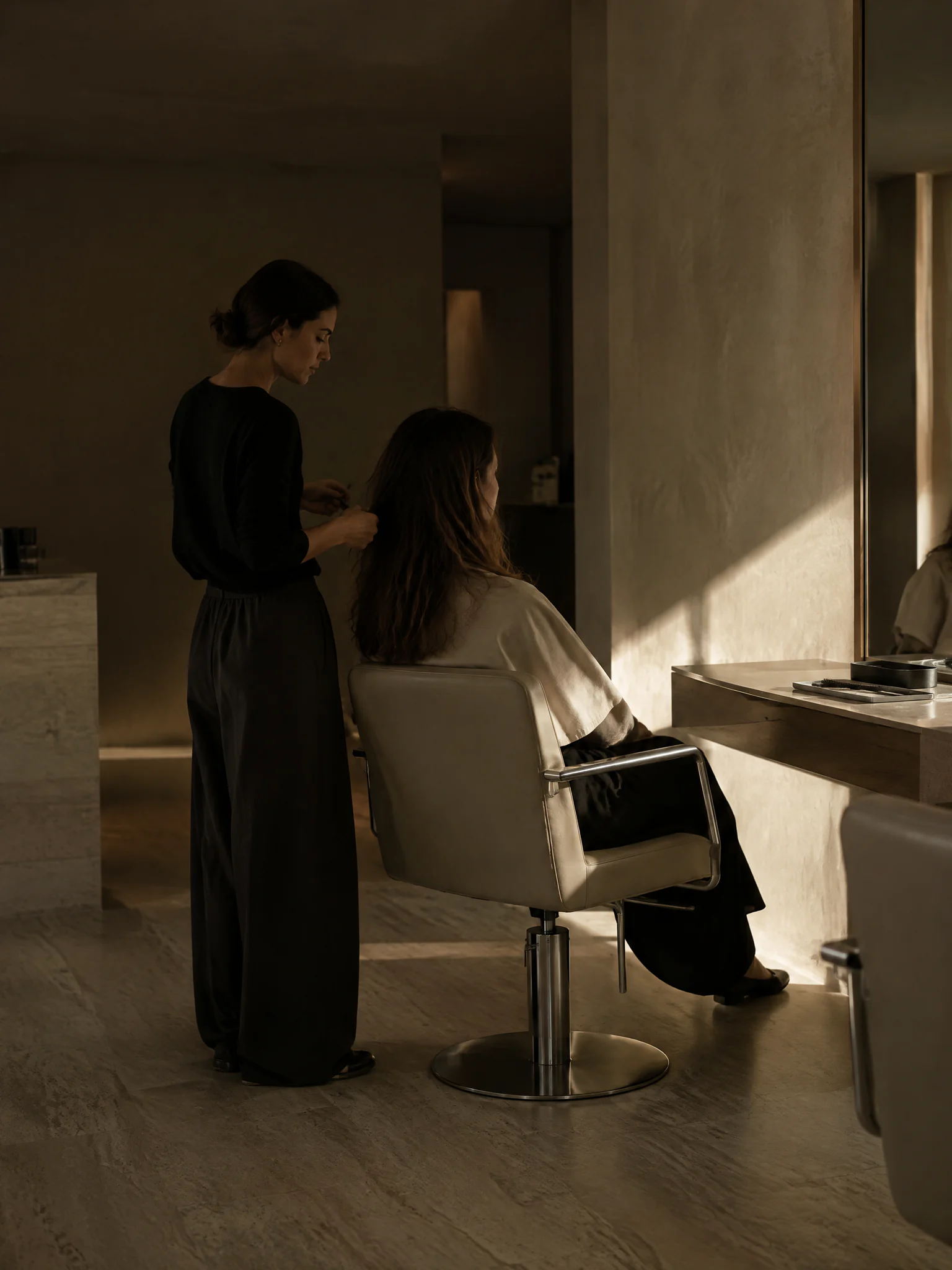 Mulher de preto a escovar o cabelo comprido de uma cliente num salão com pouca luz